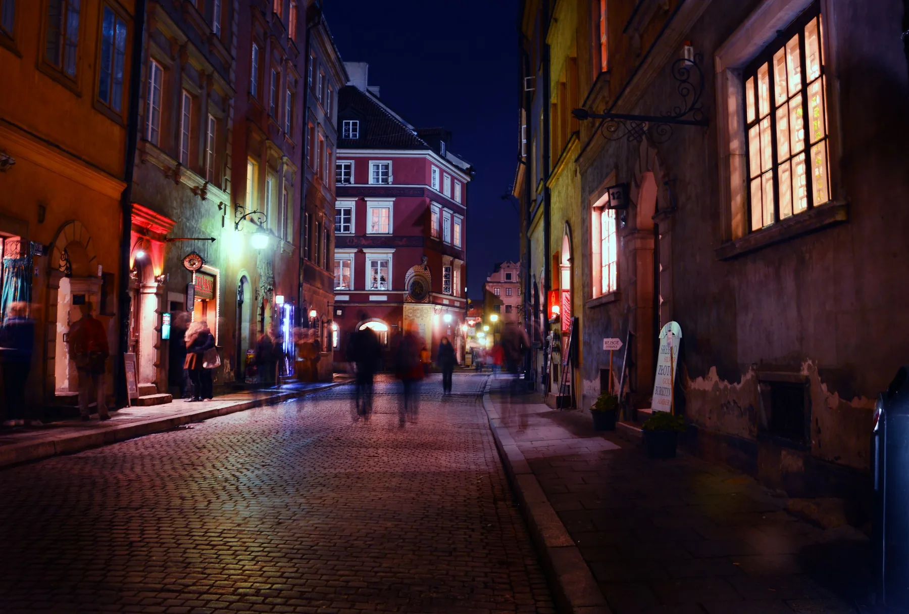 A cobbled old-town street at night with lit windows and passers-by blurred in the dusk