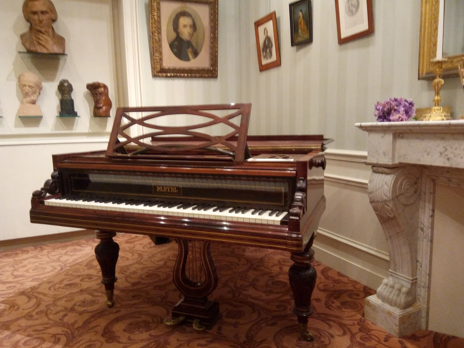 A polished Pleyel grand piano with a carved lyre pedal stand in a panelled salon, framed portraits on the wall behind