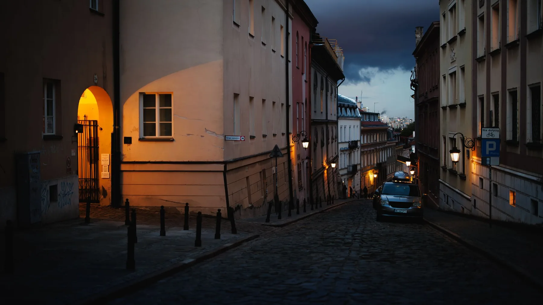 A quiet cobbled street in Warsaw's Old Town at twilight, with a lit archway on the left